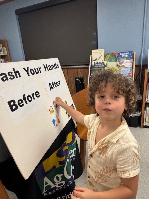 Child placing stickers on a “Wash Your Hands Before After” board in a library setting.